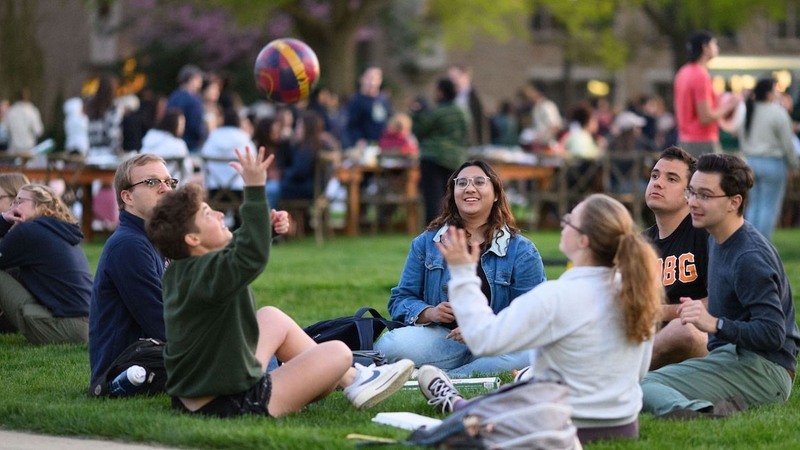 A group of college students relax on a grassy quad on a sunny afternoon. Some students chat and laugh while others watch a student in a green sweatshirt toss a ball in the air.